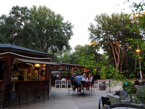 Un área de comedor al aire libre con mesas y sillas de madera, rodeada de árboles y una zona de bar con luces colgantes.