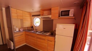 A compact kitchen with wooden cabinets, a gas stove, a microwave, and a refrigerator. - Camping L'Art De Vivre (Chateauneuf-du-Pape)