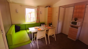 A compact kitchen with a dining table, chairs, a microwave, and a window with curtains. - Camping L'Art De Vivre (Chateauneuf-du-Pape)