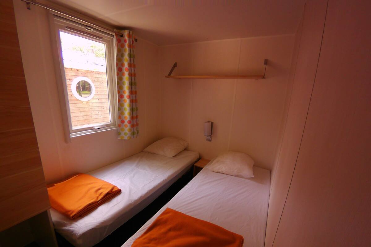 A small, compact room with two single beds, a window with a curtain, and a wooden shelf above the beds.