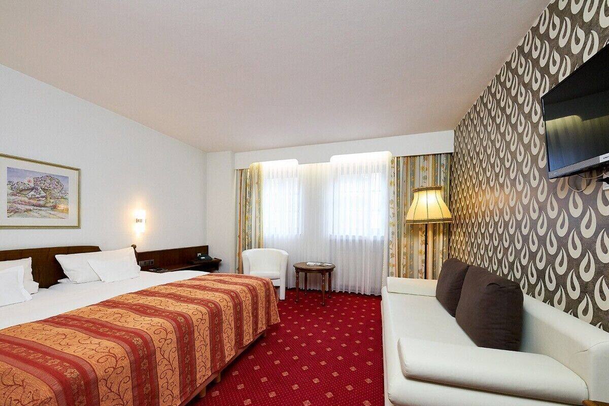 A hotel room with a large bed, a sofa, a small table, a chair, and a TV mounted on the wall.
