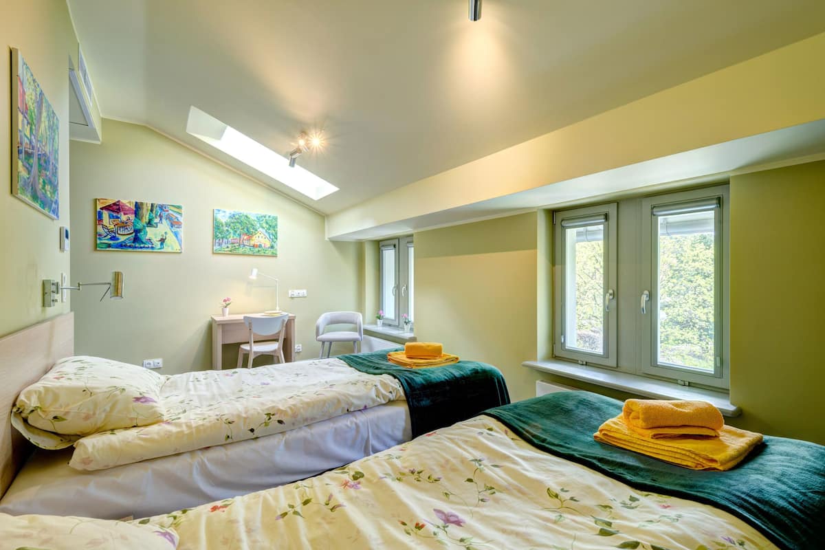 A bedroom with two beds, a desk, and a chair. There are paintings on the wall, a window with blinds, and a skylight.