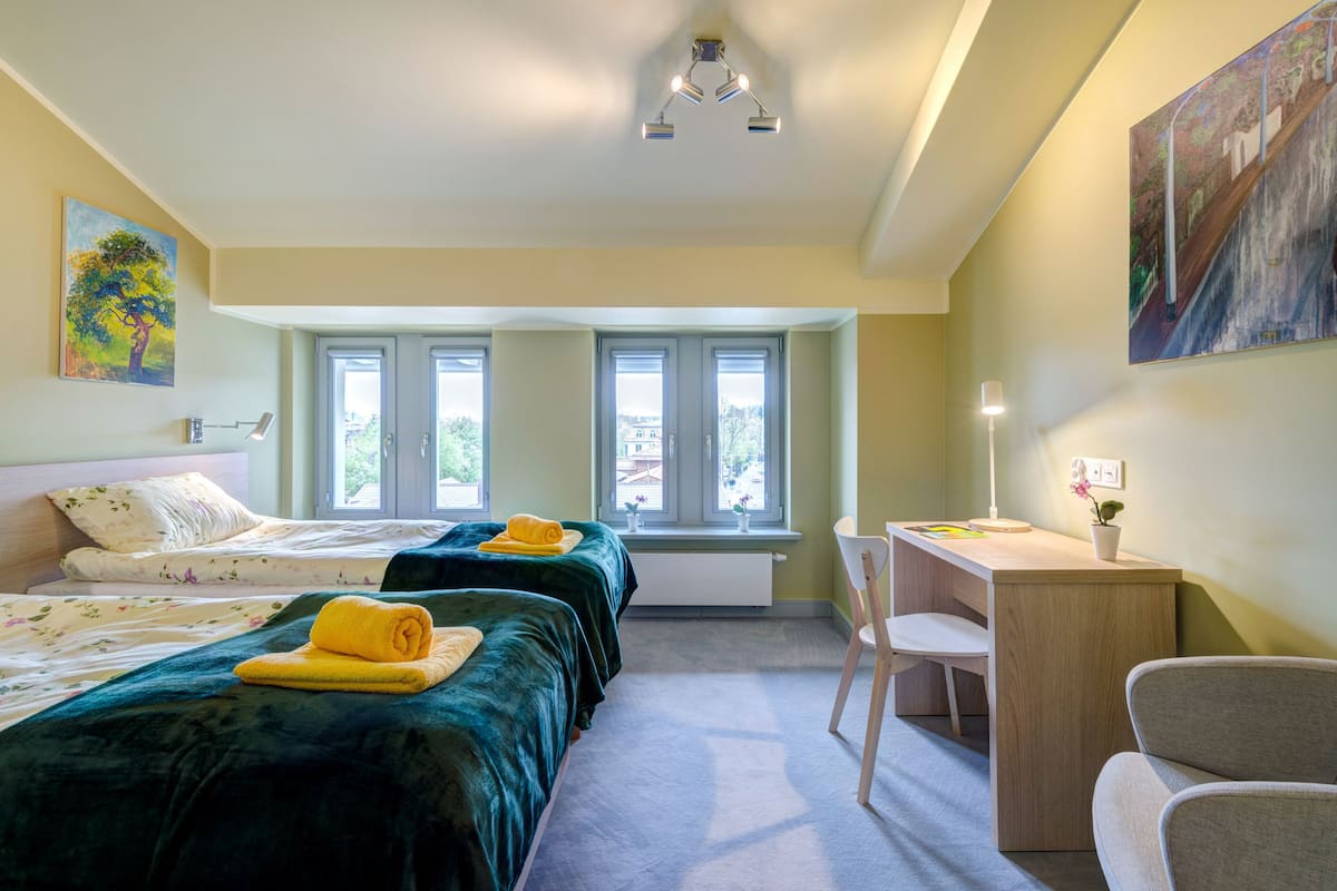 A hotel room with two beds, a desk, and a chair. There are windows with a view of greenery outside.