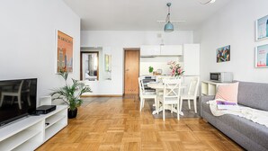 Un salon moderne doté d’un téléviseur à écran plat, d’un coin repas avec une table et des chaises blanches, et d’un canapé gris.