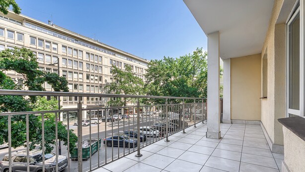 Un balcon doté d’un sol carrelé, d’une rambarde en métal et offrant une vue sur les voitures garées et un immeuble situé de l’autre côté de la rue.