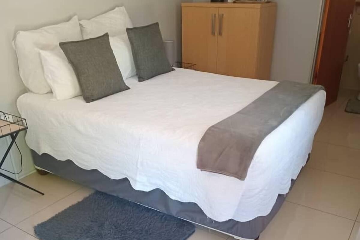 Una cama bien hecha con edredón blanco y manta gris, un armario de madera y una alfombra gris sobre suelo de baldosas.