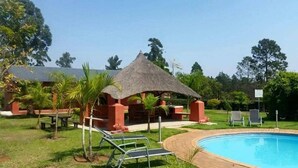 A pool area with a thatched-roof structure, lounge chairs, and a small building with a red exterior. - Brookside Lodge B&B (Matsapha)