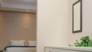 A bedroom with a bed, bedside table, and a small stool. There is a white cabinet with a mirror and a plant on top. - Brookside Lodge B&B (Matsapha)