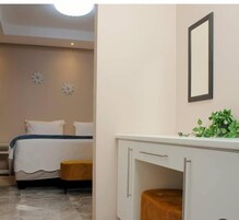 Un dormitorio con cama, mesita de noche y un pequeño taburete. Hay un armario blanco con espejo y una planta encima.