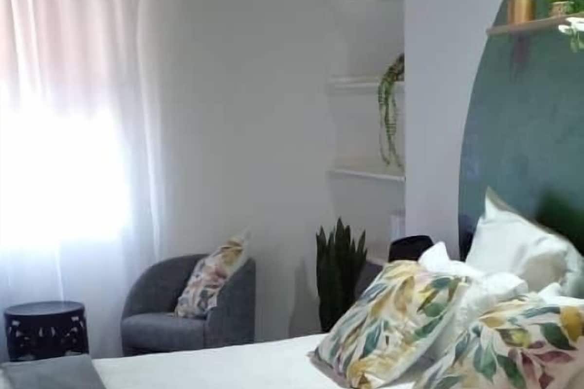 Un dormitorio con cama, una silla, una planta y un estante con objetos decorativos.