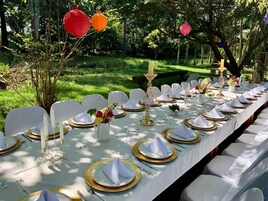 Una larga mesa de comedor al aire libre con sillas blancas, platos con detalles dorados y velas.