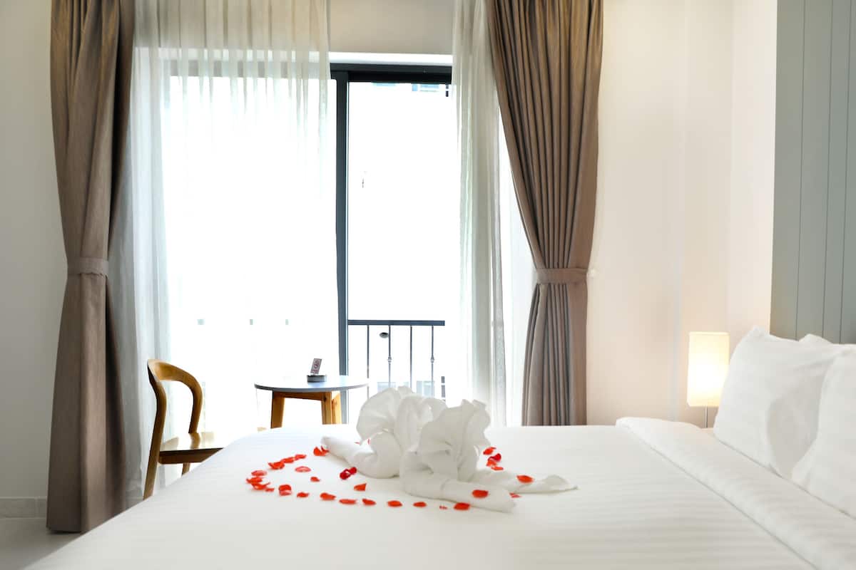 Quarto de hotel com uma cama bem arrumada, uma mesinha com uma cadeira e varanda visível pela janela.