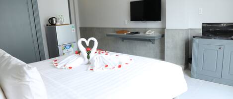 A neatly made bed with white linens and a heart-shaped decoration. A small table with a vase of flowers is beside the bed. A television is mounted on the wall above a gray shelf.