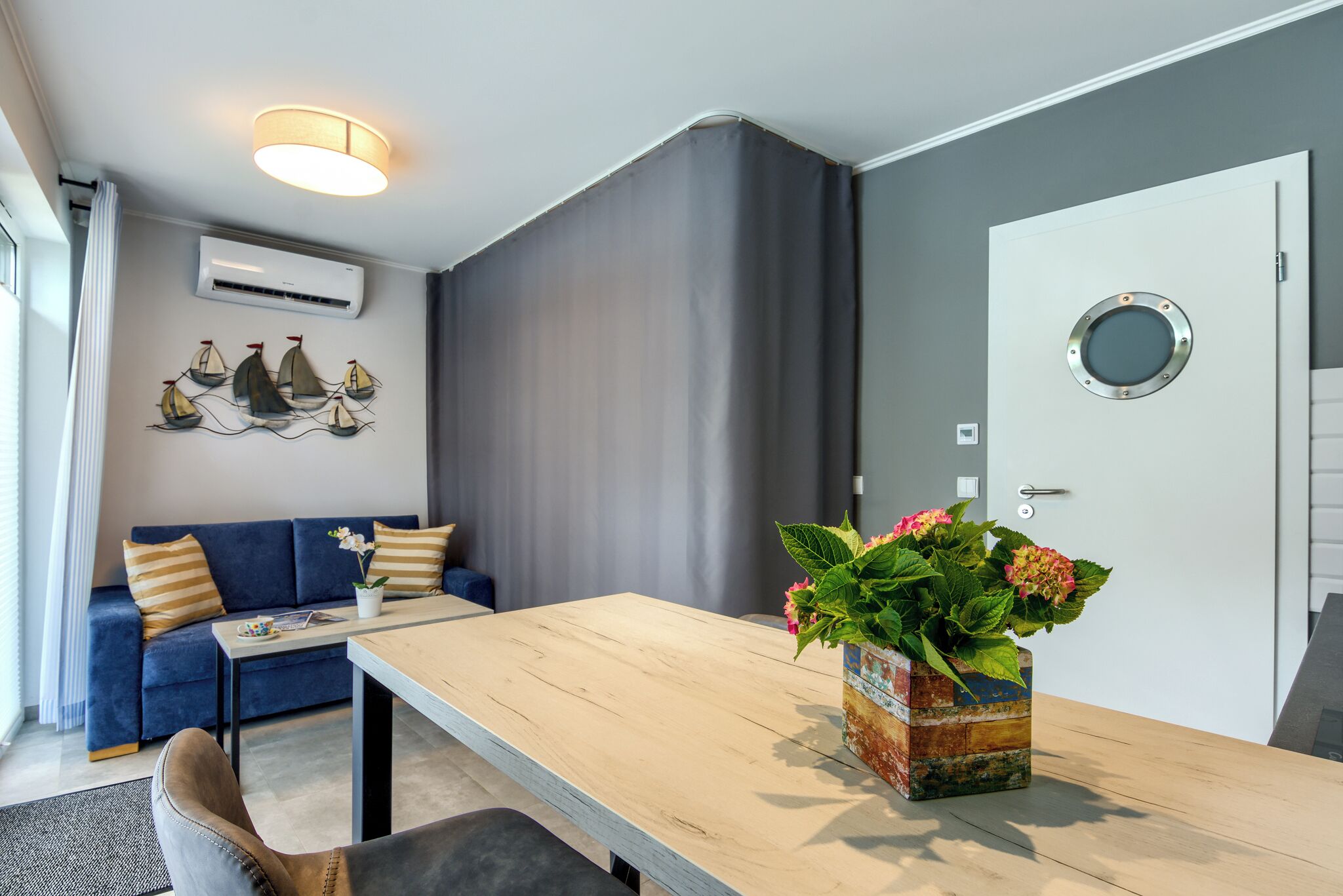 Una sala de estar moderna con un sofá, una mesa de madera, una planta en maceta y una puerta con un ventanal circular.