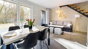 Una sala moderna con sofá, mesa de comedor y sillas. Se observa una escalera con acabado de madera y una lámpara en la pared.
