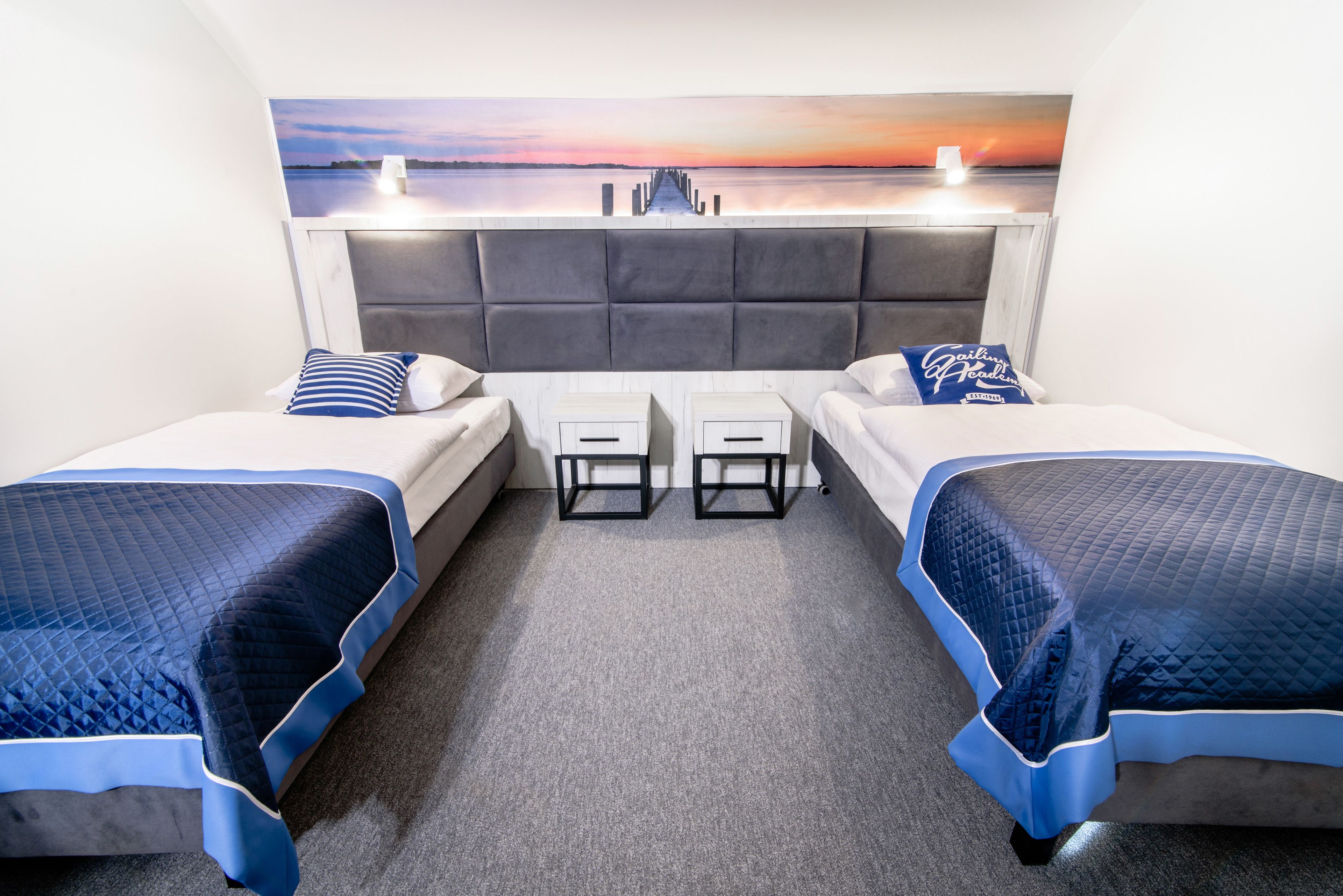 A hotel room with two beds, a grey headboard, a painting of a sunset over water, and a small table between the beds.