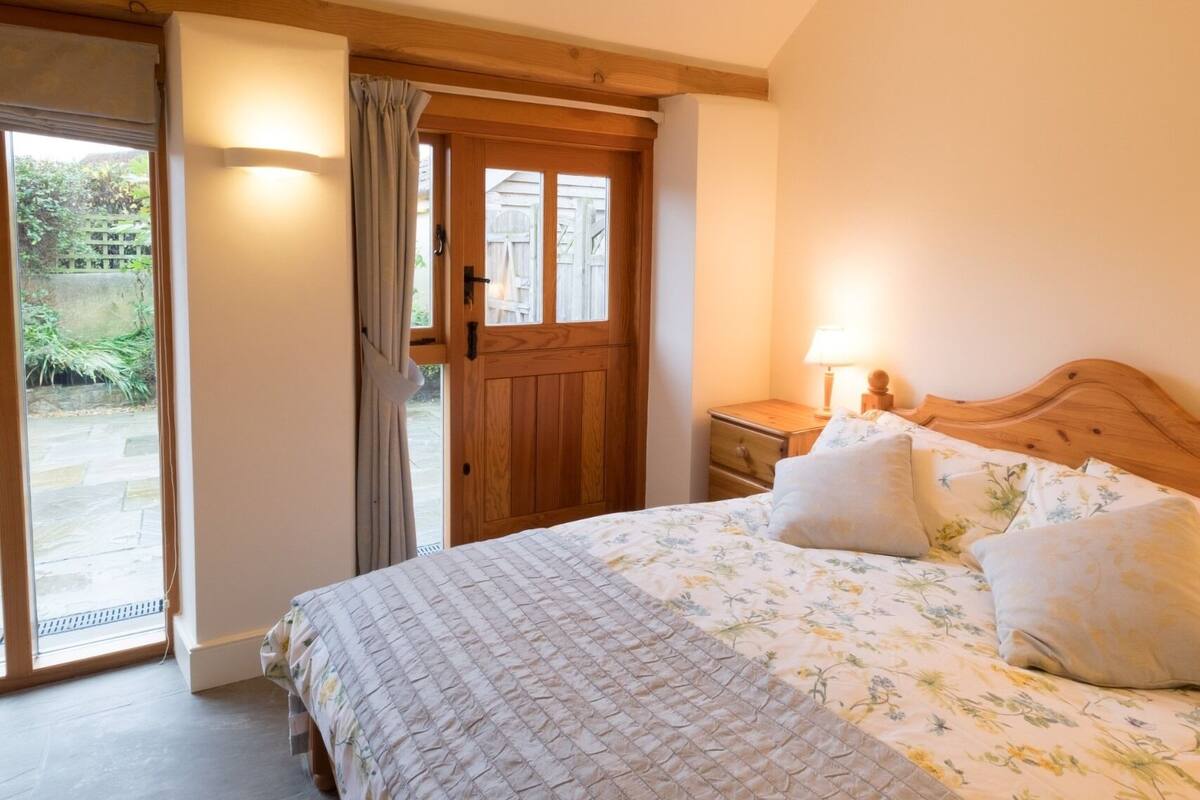 A bedroom with a wooden door, a bed with floral bedding, a nightstand with a lamp, and a view of a garden through a glass door.