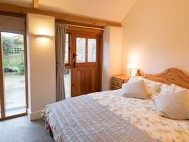 Um quarto com porta de madeira, cama com roupa de cama florida, mesa de cabeceira com abajur e vista para um jardim através de uma porta de vidro.
