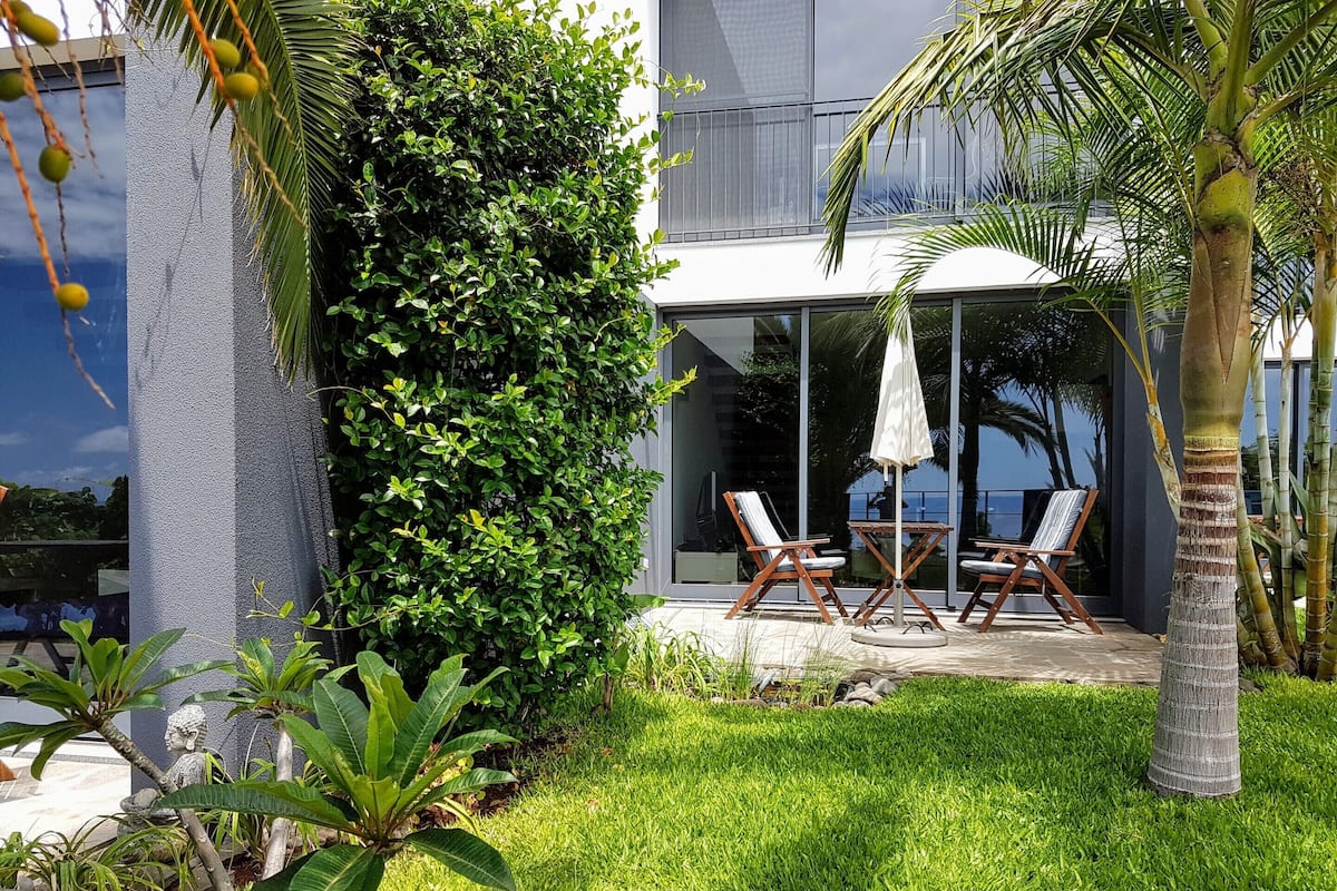 Un edificio moderno con fachada de vidrio, un patio ajardinado con dos sillones y una mesa, y un seto frondoso.