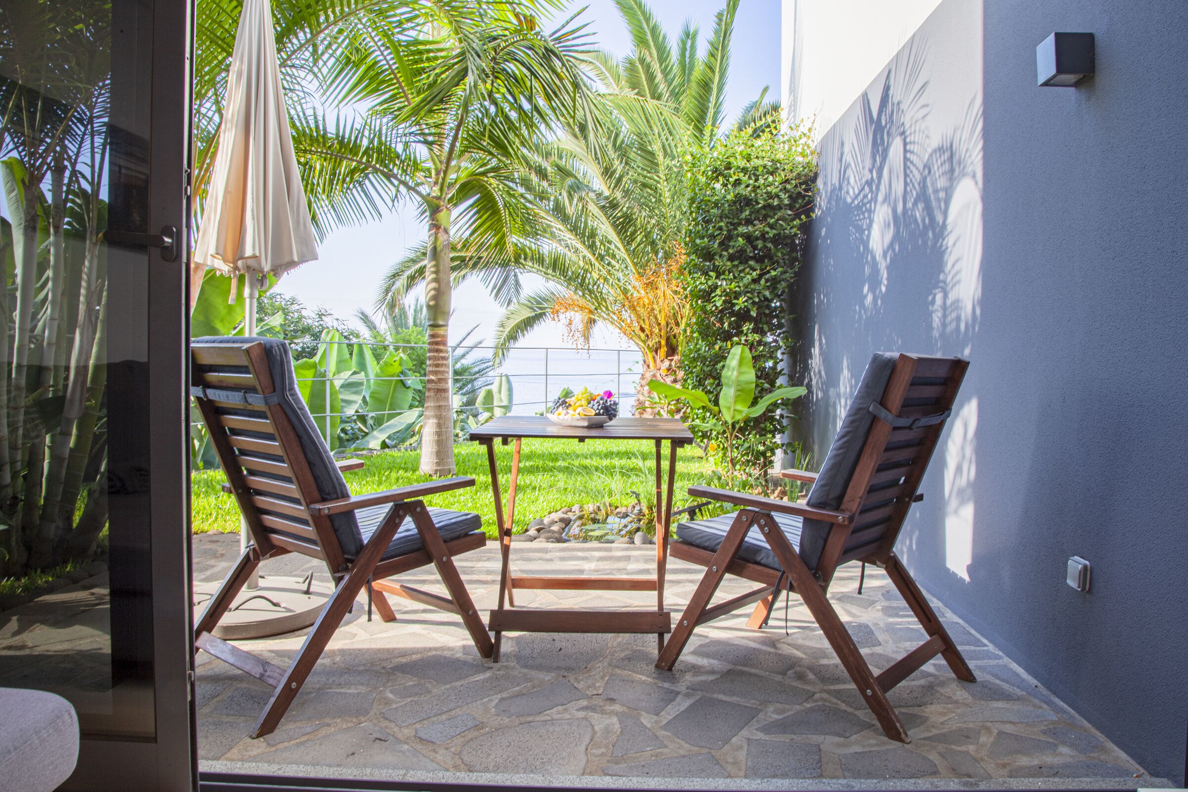 An outdoor patio with two wooden chairs and a table, surrounded by lush greenery and a palm tree.