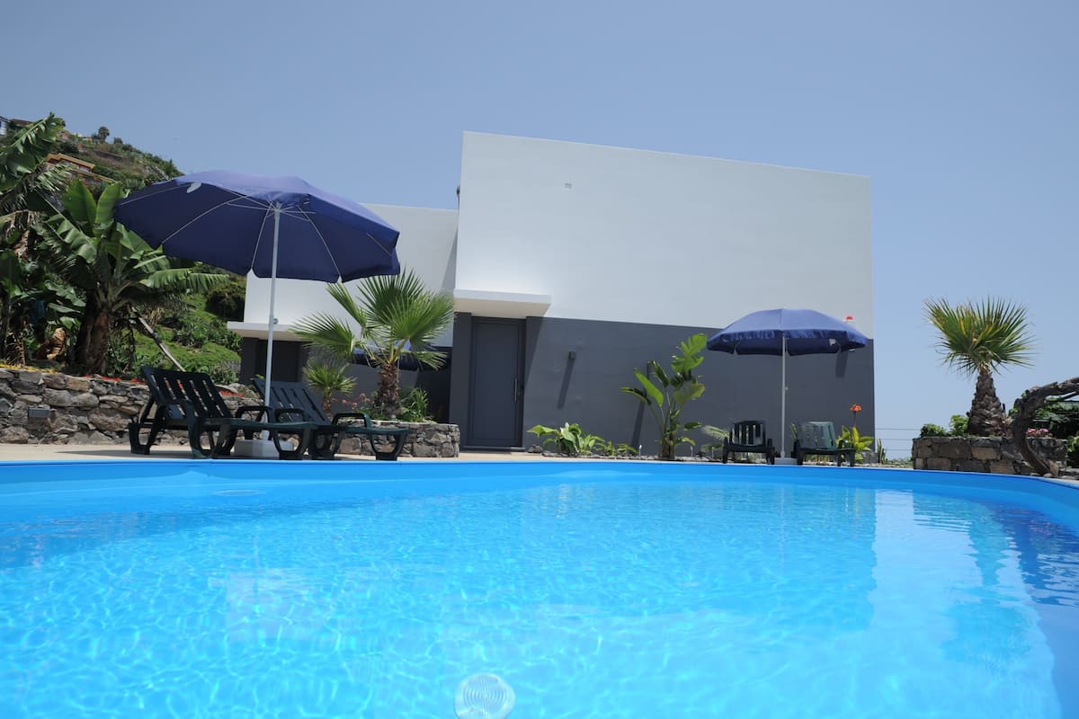 Una piscina azul cristalina con sillas de descanso y sombrillas, rodeada de plantas tropicales y un edificio moderno.