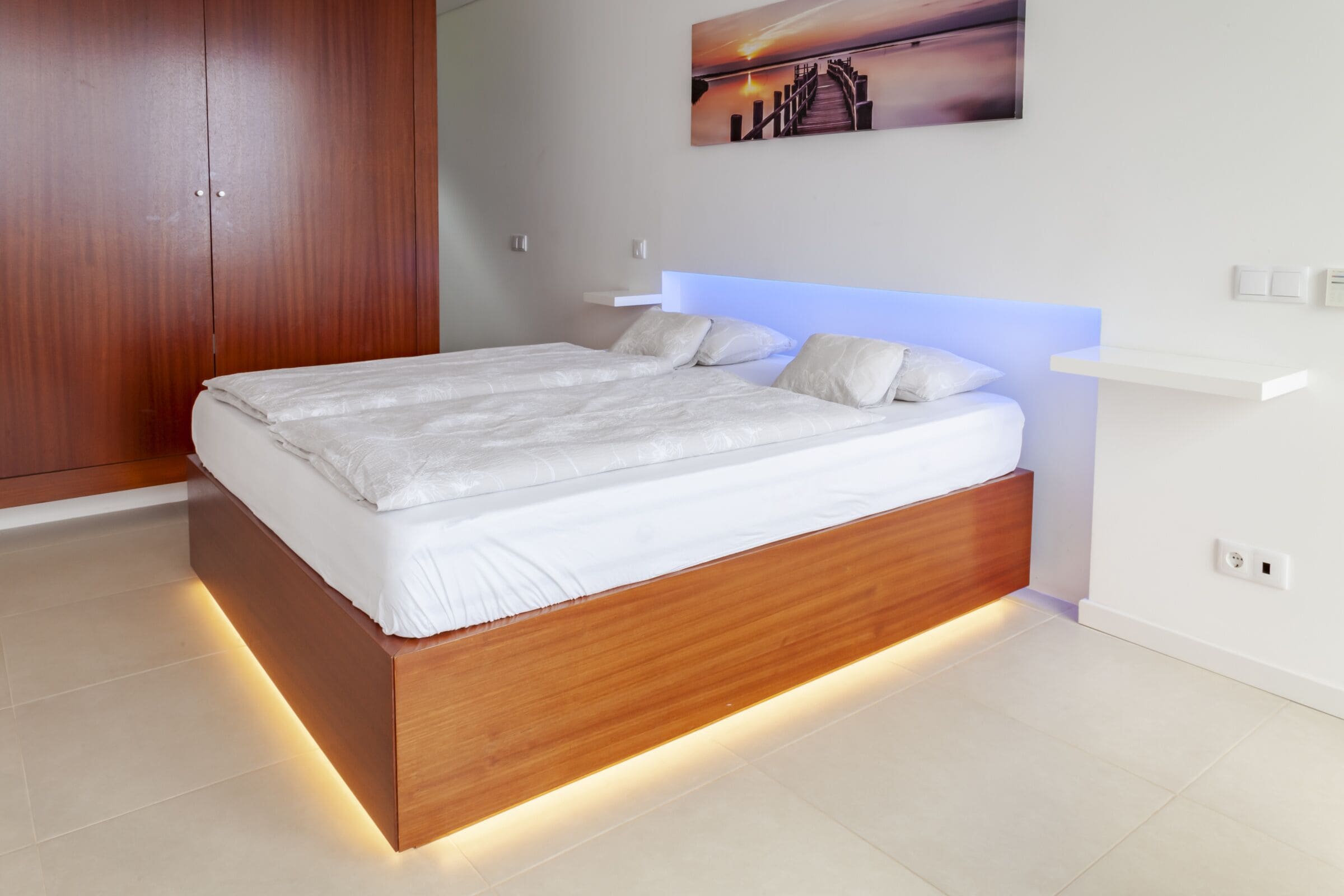 A modern bedroom with a wooden bed frame, white bedding, and a wall-mounted shelf.
