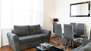 Uma sala de estar moderna com um sofá cinzento, uma mesa de centro de vidro e uma área de jantar com cadeiras e mesa.