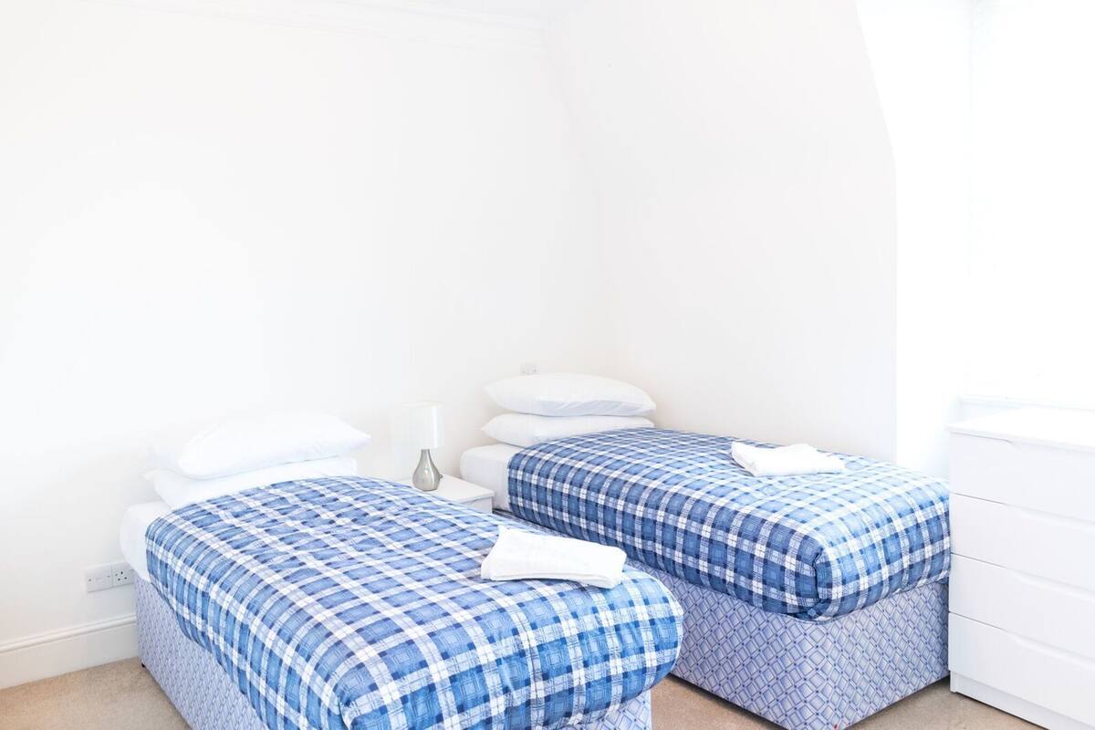 A room with two beds, each with a blue and white checkered bedspread, a bedside table with a lamp, and a white dresser.