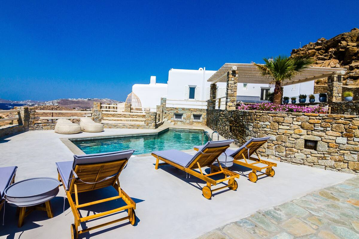 A poolside area with lounge chairs and a swimming pool, overlooking a coastal town with white buildings and a clear blue sky.