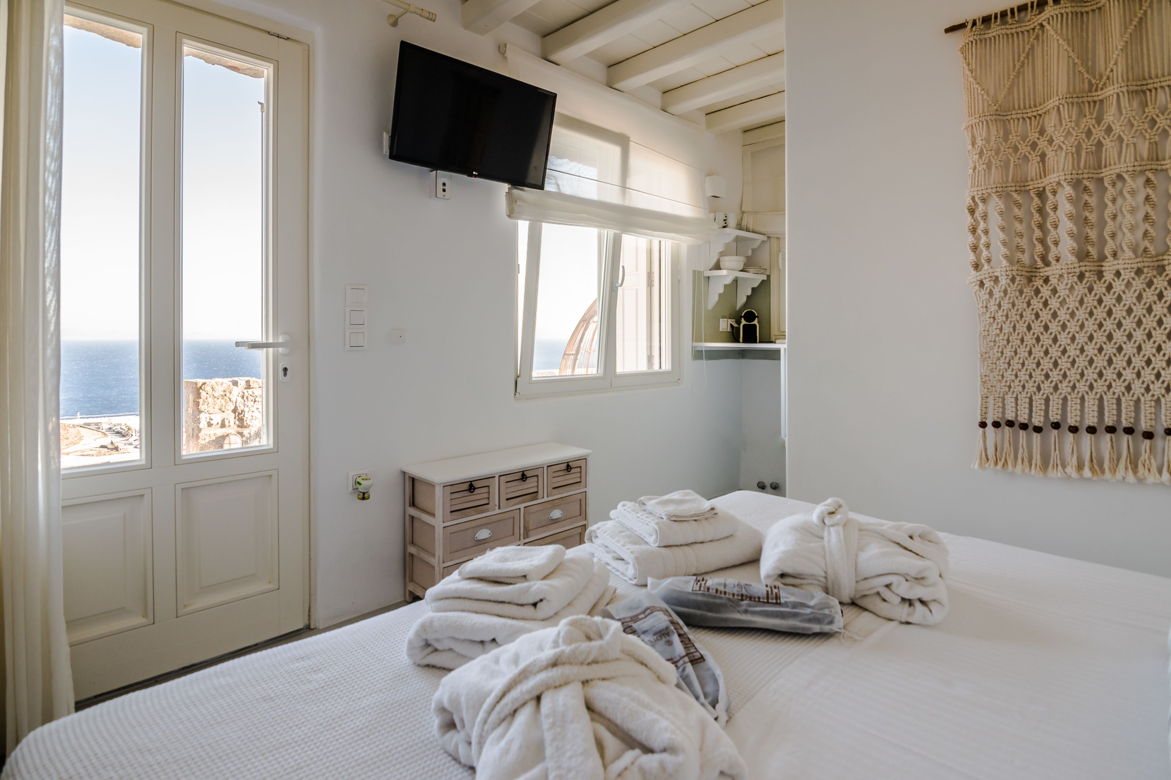 A bedroom with a bed, a TV mounted above it, a dresser, and a view of the sea through a door.