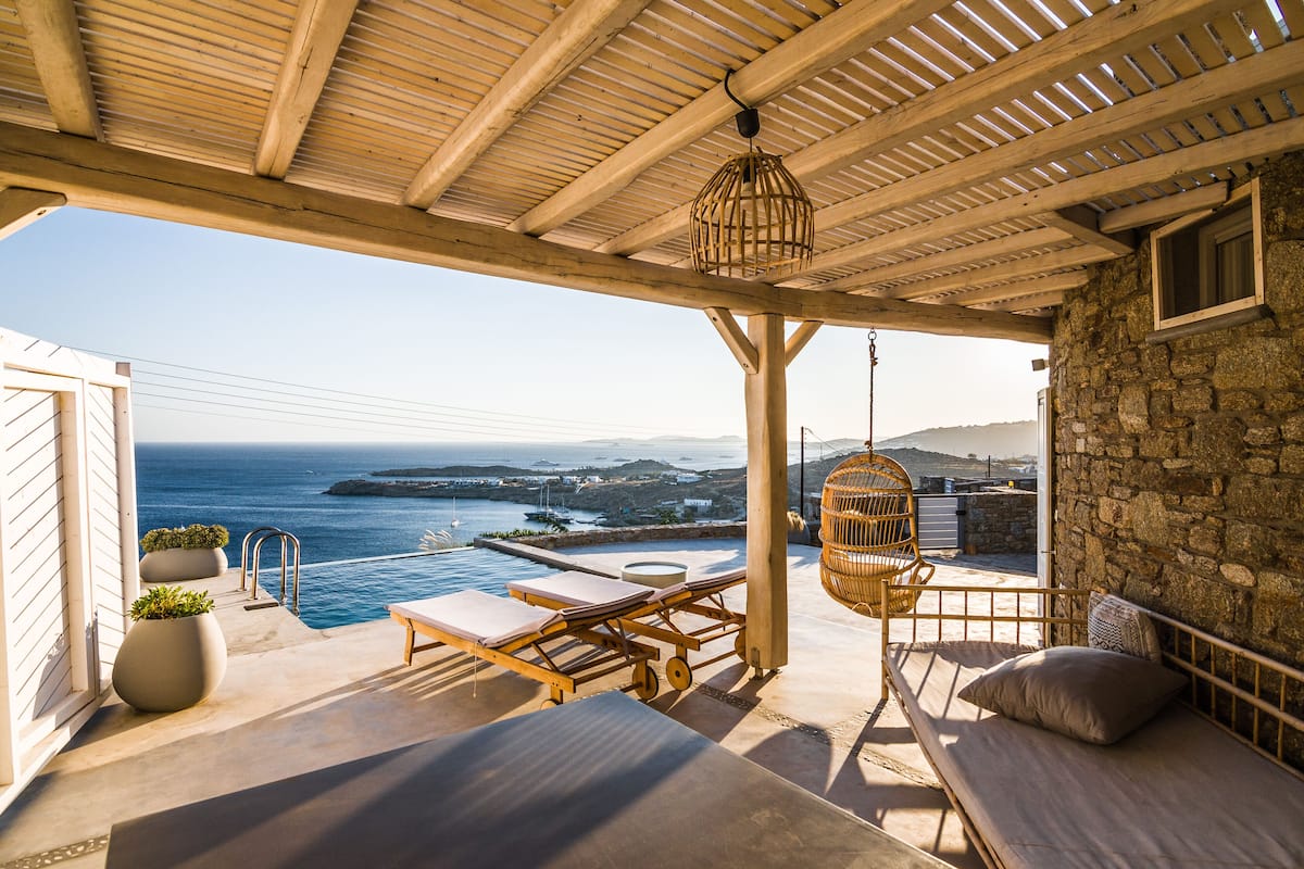 A terrace with a view of the sea, a wooden pergola, a hanging light fixture, and a stone wall.
