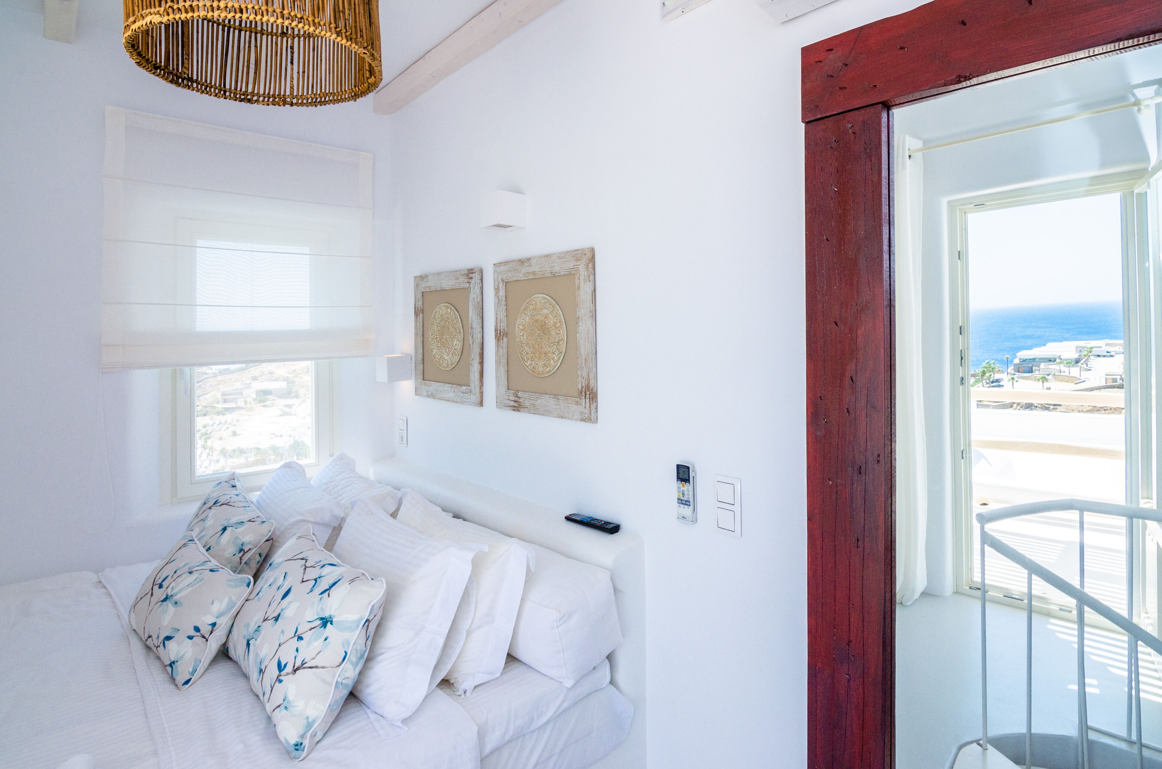 Quarto com cama, roupa de cama branca, um controle remoto e vista para a praia por uma porta aberta.