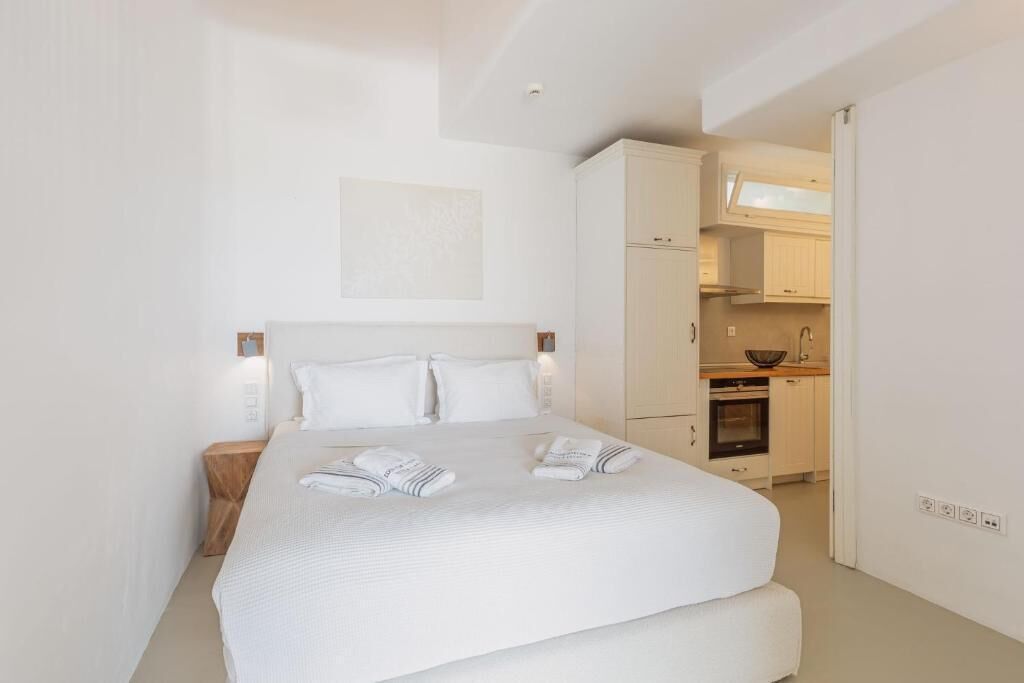 A modern hotel room with a large bed, a small bedside table, a built-in kitchen with cabinets and a stove, and a white color scheme.