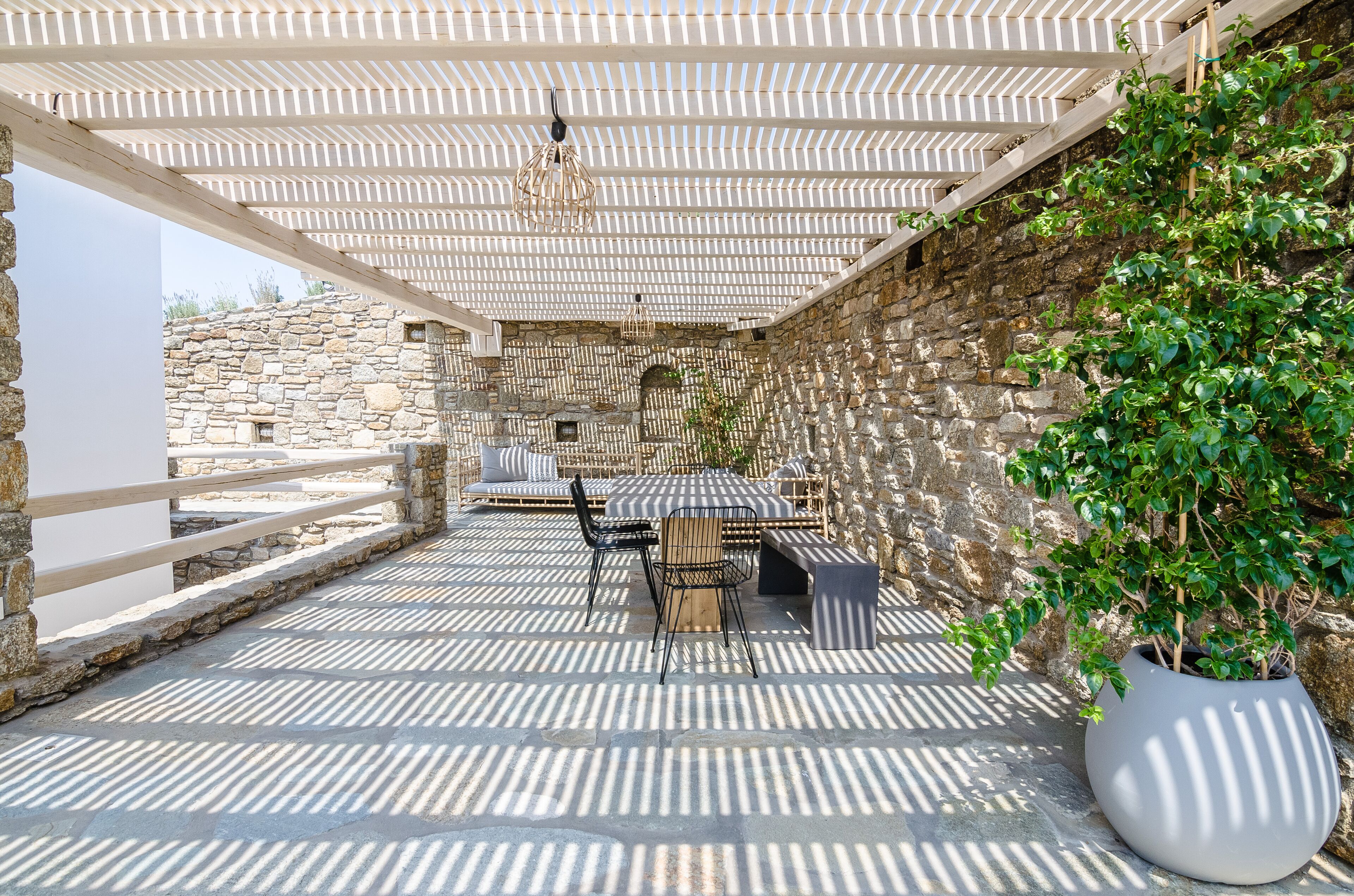 Une terrasse extérieure avec un mur en pierre, une pergola en bois et un pot de fleurs.