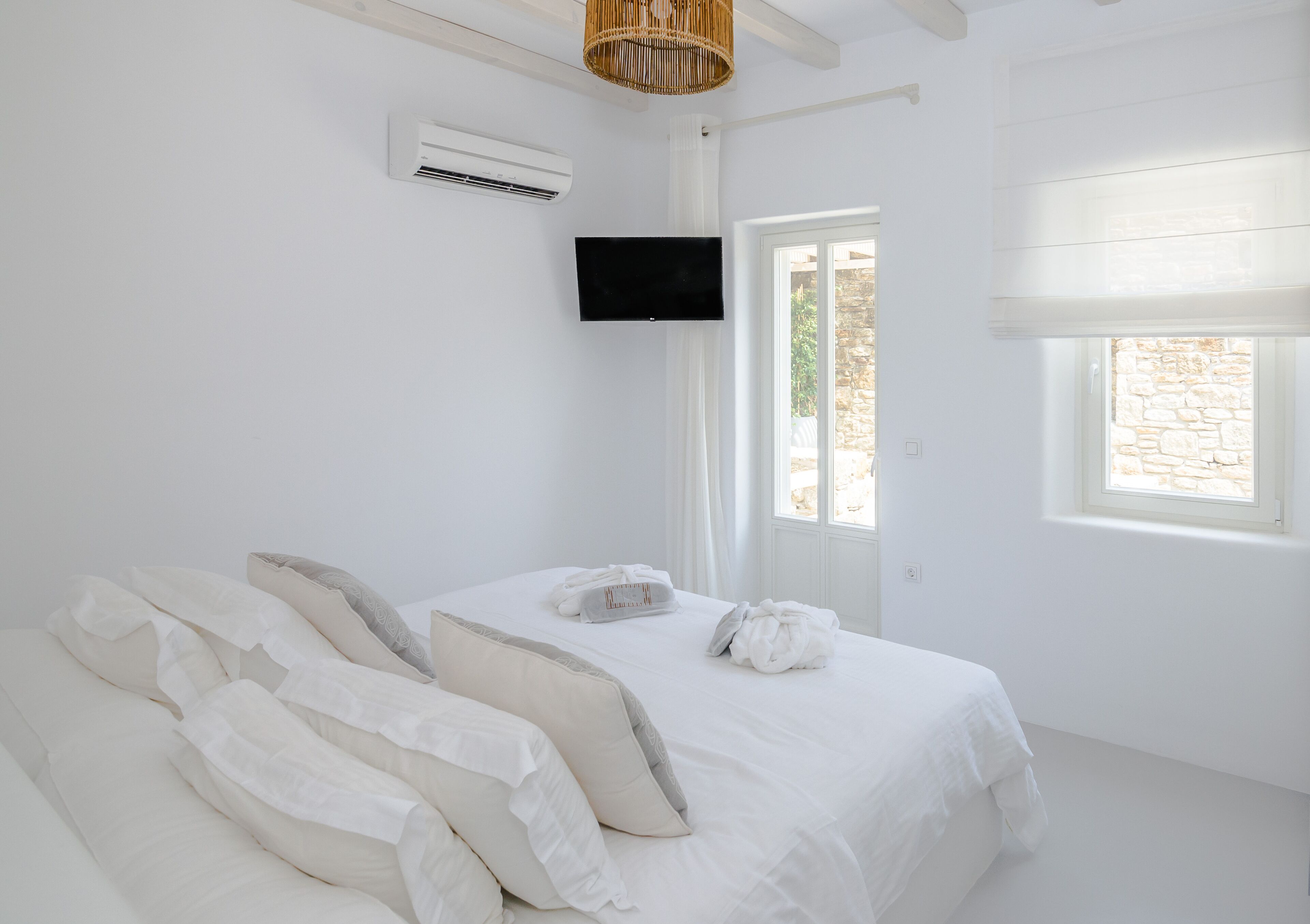 A bedroom with a bed, white bedding, a flat-screen TV mounted on the wall, and a window with a view of the outside.