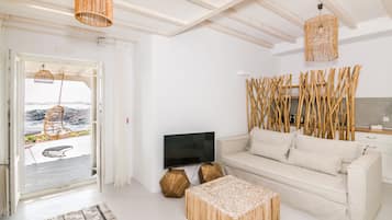 Un salon moderne avec un canapé, une table basse et un téléviseur. On peut admirer la mer à travers une porte ouverte.