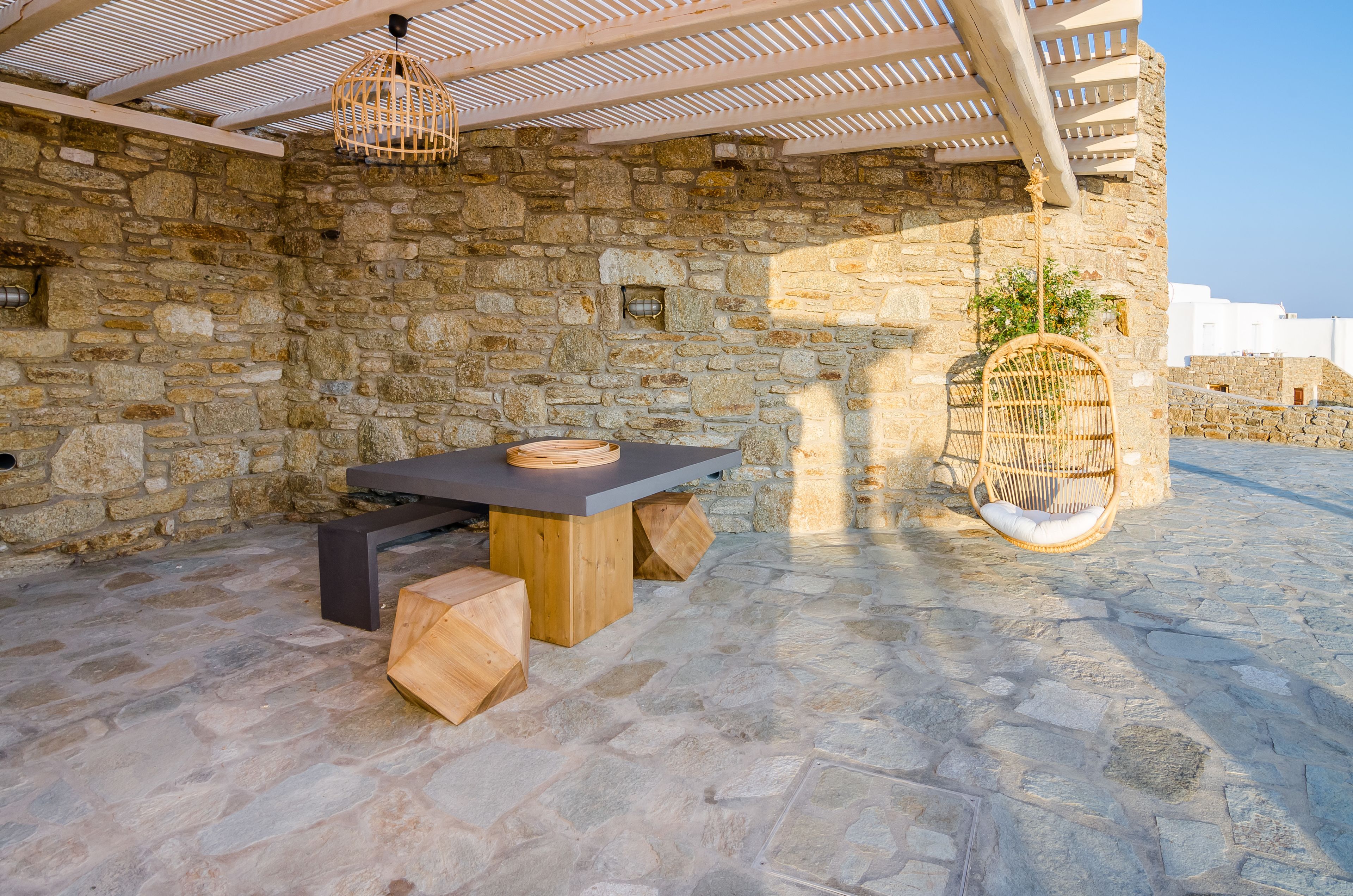 Un área de estar al aire libre con paredes de piedra, una mesa y bancos, una silla colgante de mimbre y una pérgola arriba.