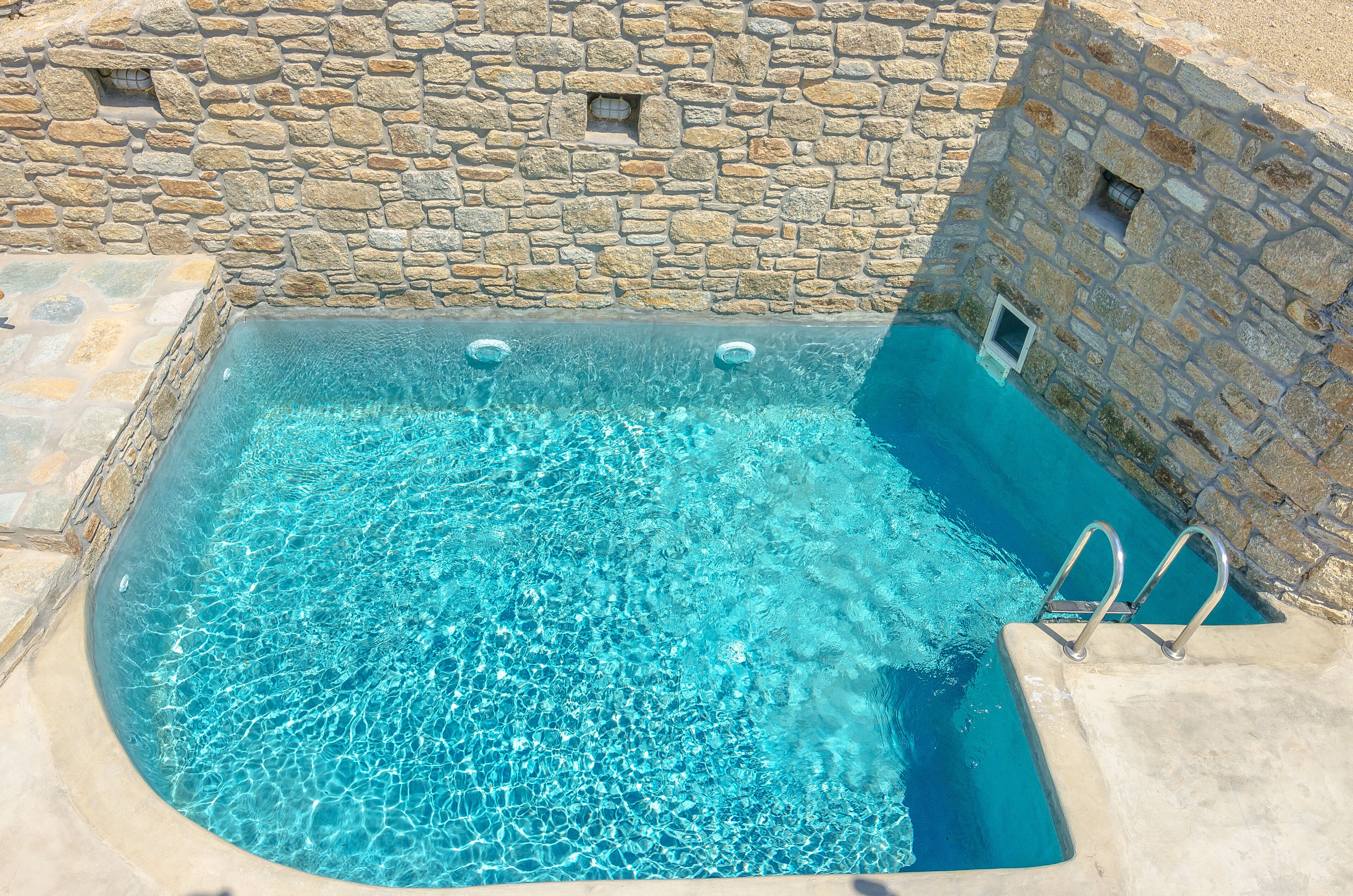 A stone-walled swimming pool with clear blue water and metal ladders.