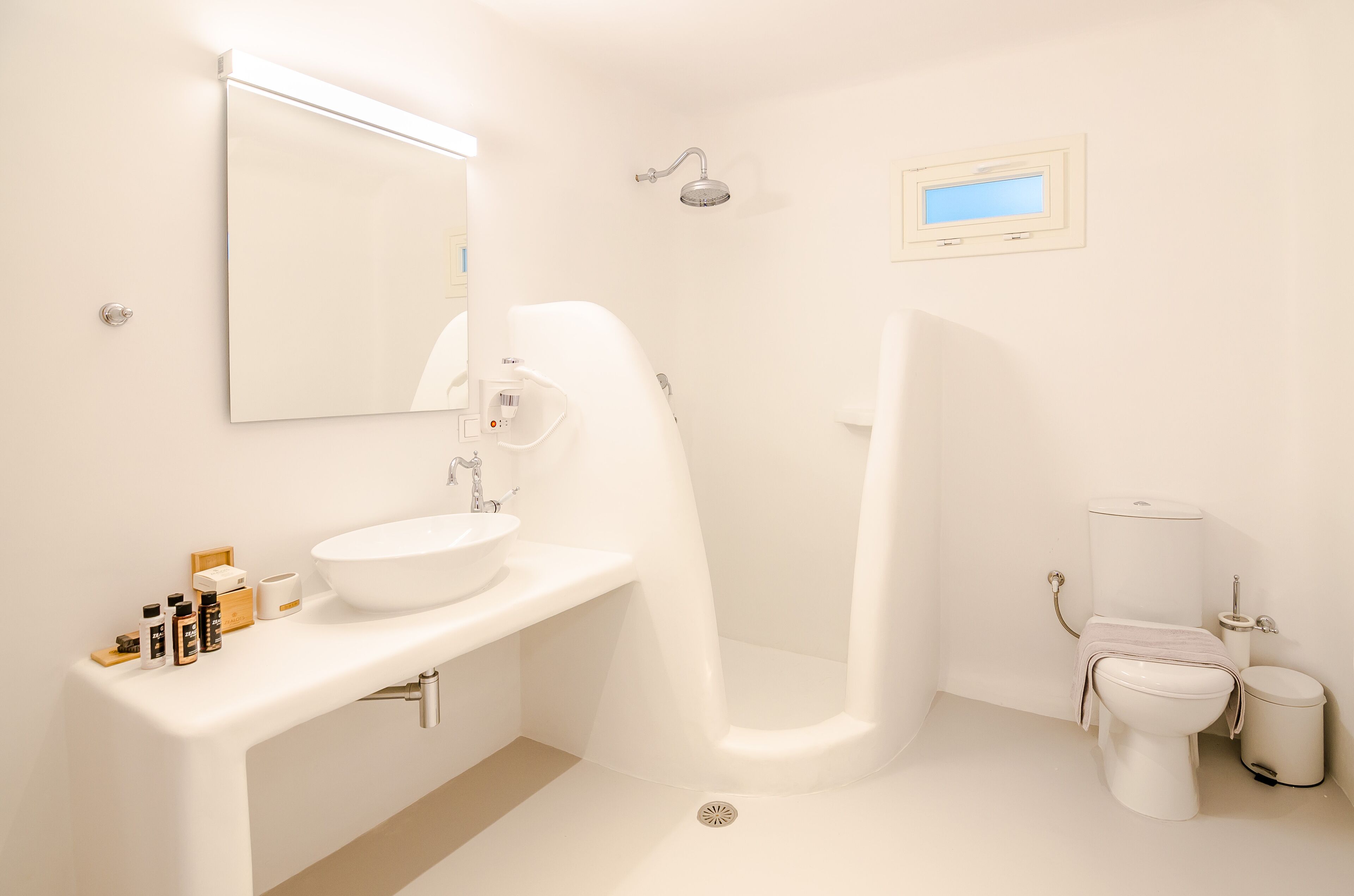 A modern bathroom with a white sink, mirror, shower, toilet, and a small window.