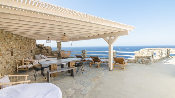 A covered outdoor seating area with stone walls, a wooden table, and lounge chairs overlooking the sea.