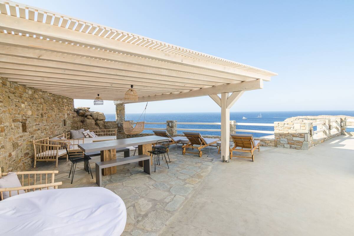 A covered outdoor seating area with stone walls, a wooden table, and lounge chairs overlooking the sea.