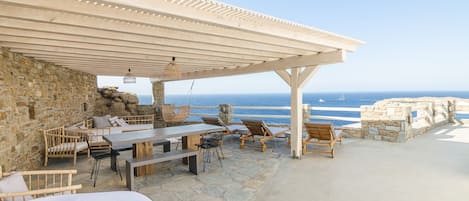 A covered outdoor seating area with stone walls, a wooden table, and lounge chairs overlooking the sea.