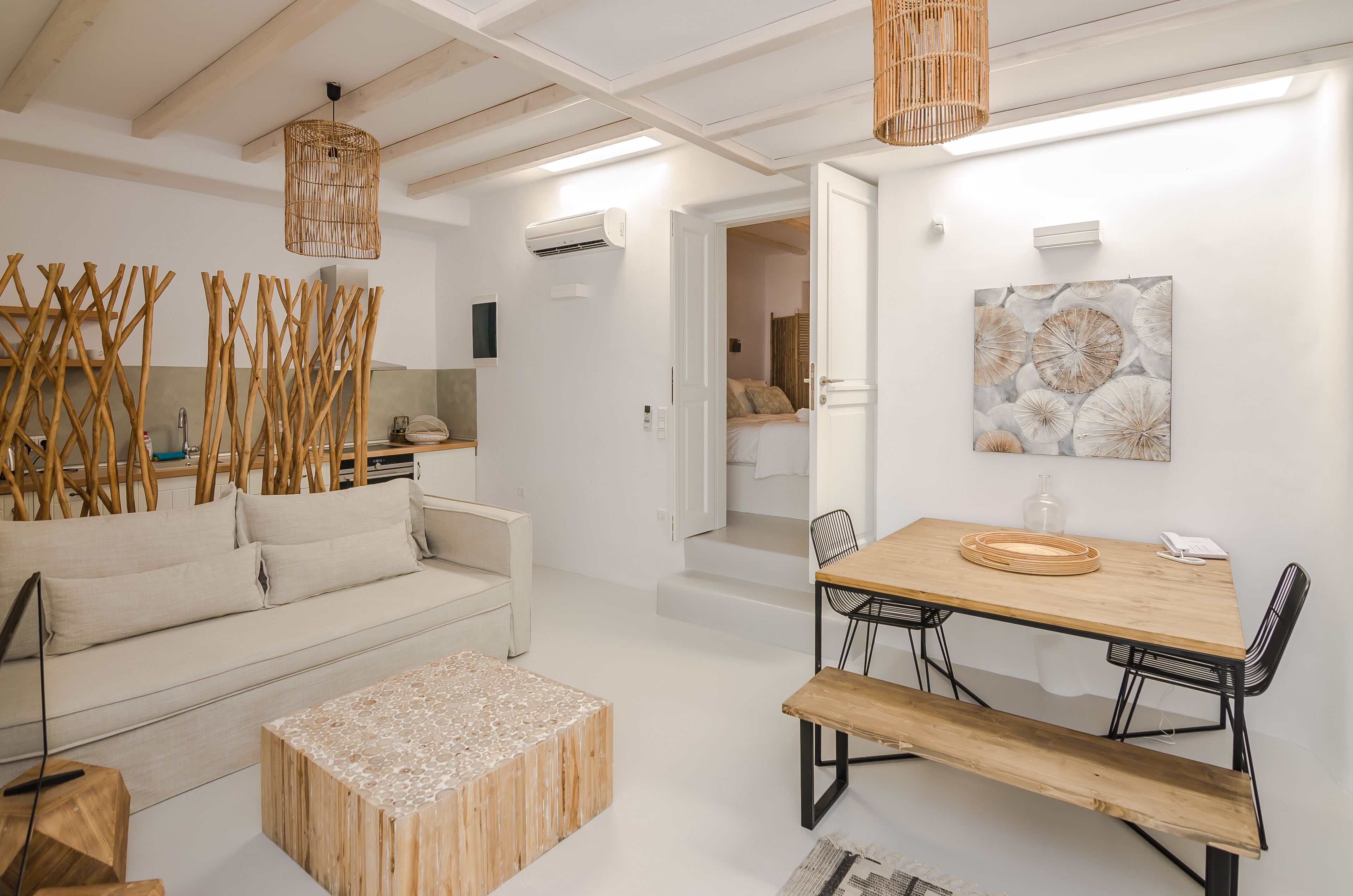 A modern living room with a sofa, coffee table, and dining table with chairs. There is a wicker lamp, an air conditioning unit, and a painting on the wall.