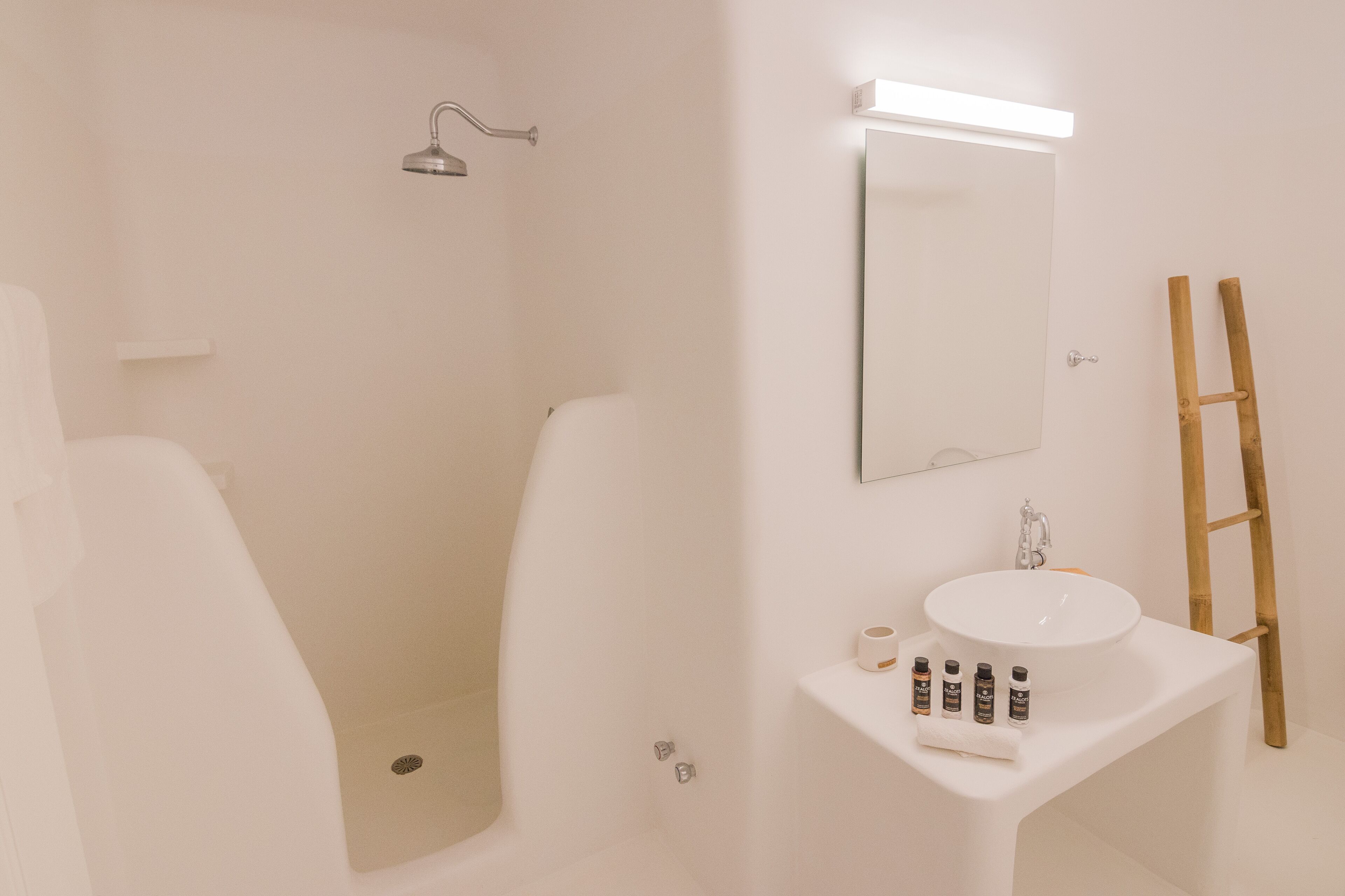 A bathroom with a white sink, mirror, and shower. There are toiletries on the sink and a ladder against the wall.