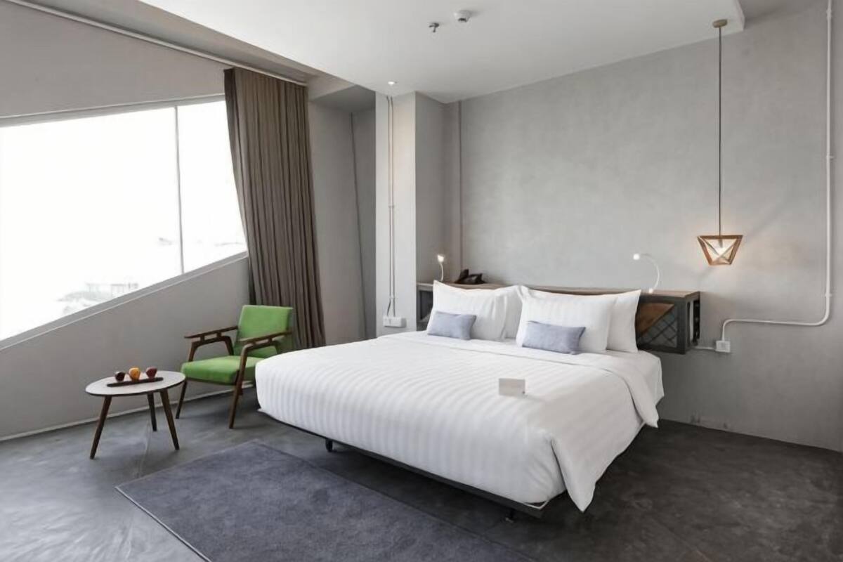 A modern hotel room with a large bed, a small table with fruits, a green chair, and a view of the city.