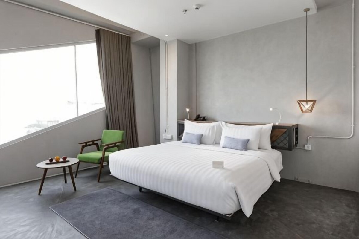 A modern hotel room with a large bed, a small table with fruits, a green chair, and a view of the city.