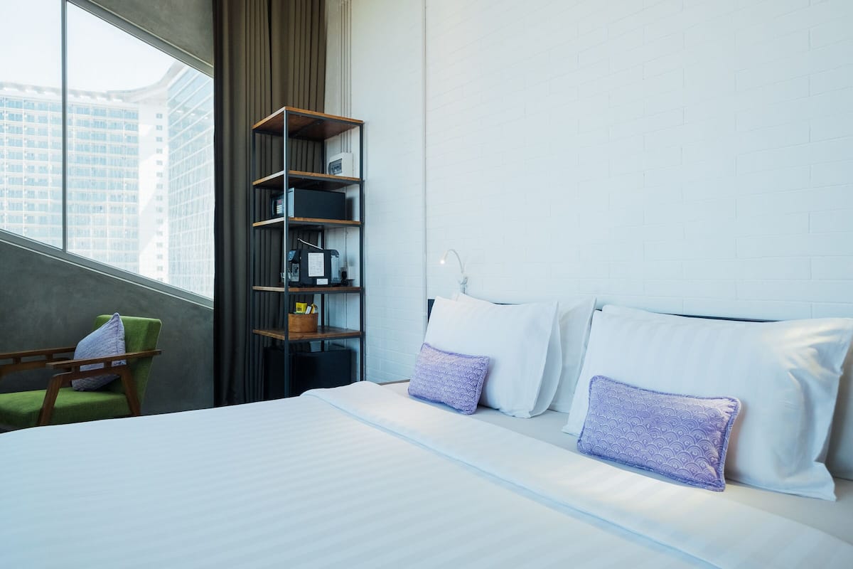 A hotel room with a bed, a green chair, a bookshelf, and a view of tall buildings.