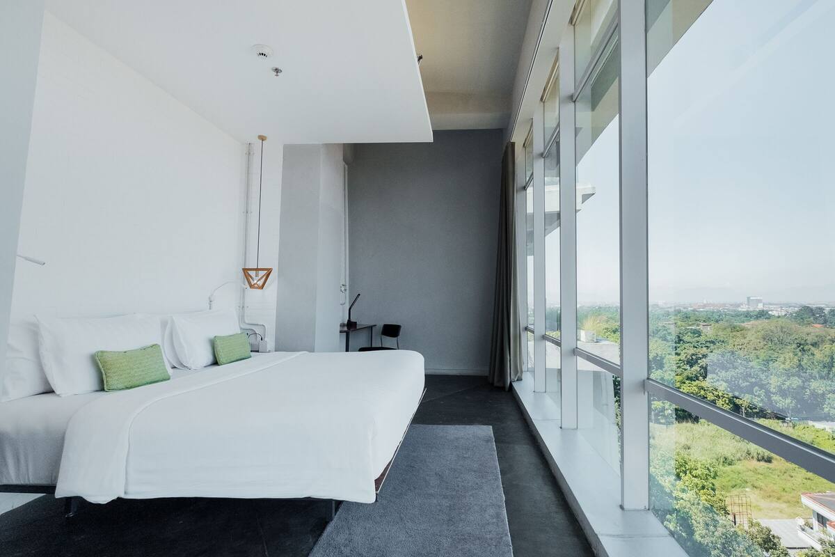 A modern hotel room with a large bed, a small desk, and a view of a green landscape through large windows.