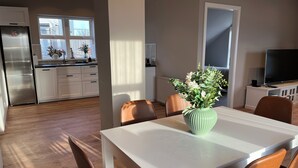 A modern kitchen with white cabinets, a stainless steel refrigerator, and a dining area with a white table and brown chairs. - Hvammur Apartments (Höfn)