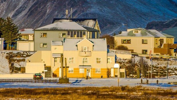 A building with a flat roof and a central entrance, surrounded by a snowy landscape and a mountain in the background. - Hvammur Apartments (Höfn)